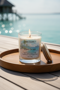 A lit Candle Elegance Island Escape scented candle in a clear glass jar sits on a round wooden tray, with its wooden lid leaning beside it. The background shows a blurred tropical ocean and overwater bungalows, indicating a beach or resort setting.