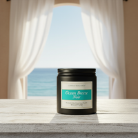 A black jar candle labeled 'Ocean Breeze Noir' by Candle Elegance sits on a light wooden surface in front of a window with white curtains and an ocean view.