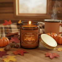 A lit amber glass jar candle labeled 'Pumpkin Spice' sits on a wooden table surrounded by fall leaves, acorns, cinnamon sticks, small pumpkins, and a mug with steam in a cozy indoor setting.