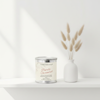 A Candle Elegance 'Purely Unadventured' soy coconut candle in a silver tin with a white wax insert, styled on a white shelf next to a white ceramic vase holding dried pampas grass.