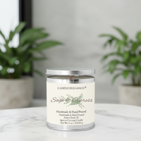 A silver metal jar candle with a white label reading 'Candle Elegance Sage & Cypress' sits on a light marble surface with blurred green potted plants in the background.