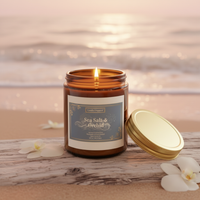 An amber glass jar candle labeled 'Sea Salt & Orchid' by Candy Dragonfly, lit with a small flame, set on a beach log with a gold lid beside it and white orchids in the foreground, against a blurred ocean sunset background.