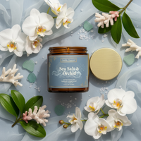 A jar candle labeled 'Sea Salt & Orchid' by Candle Elegance, shown with its gold lid beside it on a light blue fabric backdrop, surrounded by white orchids, green leaves, and small coral pieces.