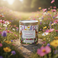 A paint can tin candle labeled 'Candle Elegance Sunwashed' sits on a stone surface in a sunlit field of pink, purple, and yellow flowers. The label features floral and butterfly artwork, and the background is a soft-focus meadow with warm sunlight.