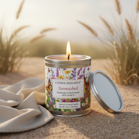 A lit Candle Elegance Sunwashed scented candle in a silver metal tin sits on a sandy surface with a beige cloth beside it and blurred grass in the background. The lid rests open to the right.