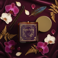 A square amber glass candle jar labeled 'Vanilla Orchid' by Candle Elegance, styled on deep burgundy satin with purple orchids, white orchids, vanilla beans, a feather, gold fern leaves, and a gold lid beside it.