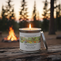 A lit candle in a silver metal tin sits on a rustic wooden surface outdoors at dusk, with a blurred campfire and trees in the background. The label reads Candle Elegance Wildflowers.