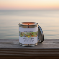 A lit metal tin candle with its lid leaning against it sits on a wooden railing with a blurred ocean sunset in the background. The label reads 'Candle Elegance' and 'Wild Flowers'.