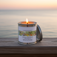 A lit tin candle labeled Candle Elegance Wildflowers sits on a wooden deck railing with a blurred ocean sunset background.