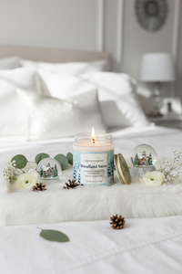 A lit candle in a clear glass jar sits on a white bed runner in a softly styled bedroom. The candle label reads 'colortraject Woodland Snow'.